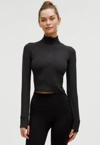 Langærmet sort træningstrøje med høj krave, lynlås foran, ribbet tekstur og tommelfingerhuller ved manchetten. Matchet med sorte højtaljede leggings.