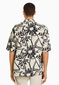 Krátký rukáv košile z krémové látky s výrazným černým květinovým vzorem, s otevřeným límcem a volným střihem, s hladkou texturou.