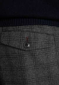 Un pantalon en plaid gris avec une texture lisse, doté d'une poche à rabat, d'un bouton noir et d'une couture d'accent rouge sur la fermeture de la poche.