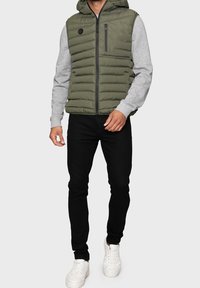 Veste sans manches olive avec manches longues grises, capuche et poche zippée noire ; associée à un pantalon slim noir et des baskets blanches.