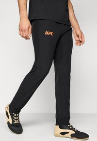 Μαύρο αθλητικό παντελόνι με πορτοκαλί λογότυπο "UFC", σχεδιασμό που στενεύει, ελαστικές μανσέτες και απαλό, ελαφρύ ύφασμα.