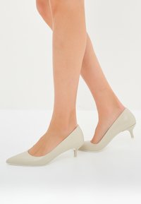 Tacchi beige a punta con tacco stiletto basso. Materiale sintetico liscio con un design pulito e moderno e un leggero arco.