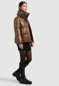 Jacheta pufosă maro cu guler înalt și textură lucioasă, asortată cu o fustă neagră cu dungi și cizme negre până la genunchi.