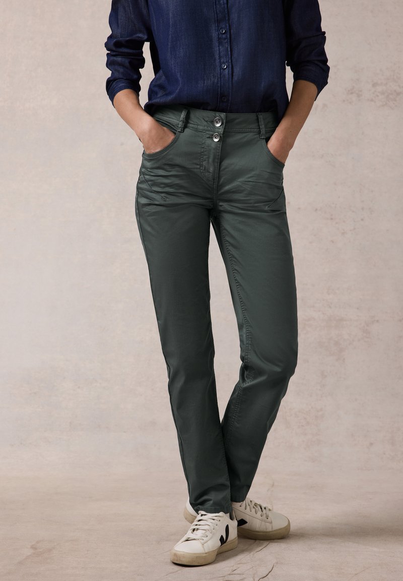 Pantalones verdes oscuros de corte ajustado, que cuentan con dos bolsillos delanteros, dos botones en la cintura y un acabado suave y texturizado. Combinados con zapatillas blancas.