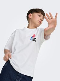 Jongen met een wit T-shirt met print en donkere spijkerbroek, die één hand naar de camera opheft tegen een effen achtergrond.