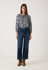 Blumige marineblaue Bluse mit langen Ballonärmeln, Schlüsselloch-Ausschnitt, kombiniert mit hoch taillierten, weit geschnittenen dunkelblauen Jeans und braunen Stiefeletten.