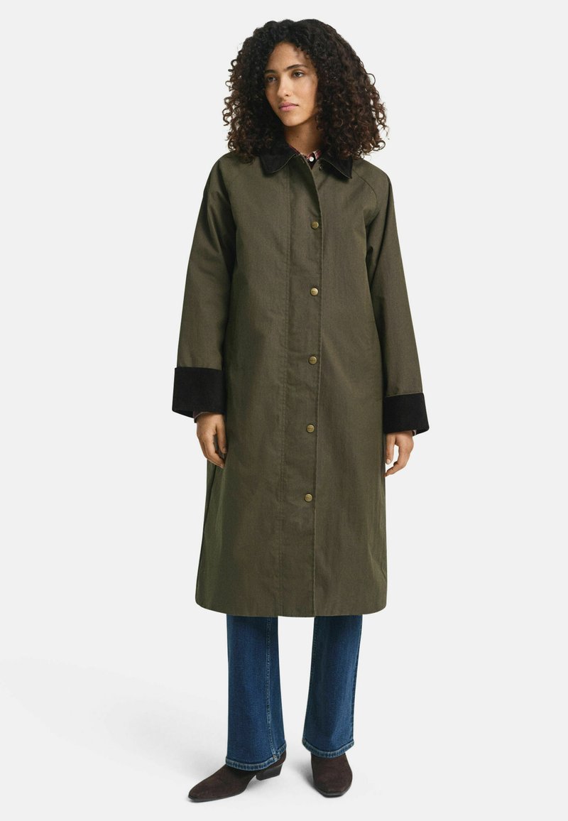 Vrouw met krullend haar, gekleed in een olijfgroene lange jas met zwarte kraag en boorden, blauwe jeans en bruine enkellaarsjes, staat rustig.