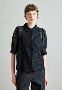 Blusa in pizzo nero con maniche corte a sbuffo, scollo con collarino e apertura frontale con bottoni. Presenta motivi floreali e texture trasparente. Abbinata a jeans scuri.