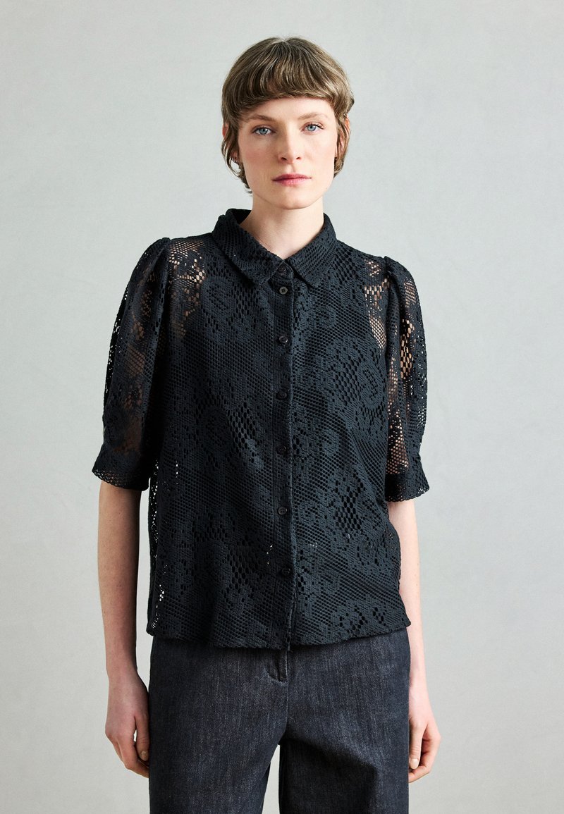 Blusa in pizzo nero con maniche corte a sbuffo, scollo con collarino e apertura frontale con bottoni. Presenta motivi floreali e texture trasparente. Abbinata a jeans scuri.