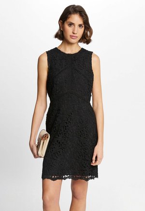 Abito in pizzo nero con motivi floreali, busto aderente e orlo a scallop. Abbinato a una borsa a tracolla neutra.