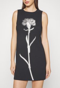Jeune femme portant une robe noire sans manches avec un grand motif pixelisé d’une rose blanche sur le devant, debout devant un fond uni.