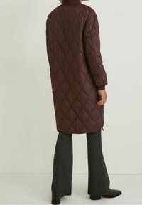 Prošívaný dlouhý kabát tmavě bordó barvy s hladkou texturou, s límcem stojacím a žebrovanými manžetami; zobrazen s kostkovanými rozšířenými kalhotami.