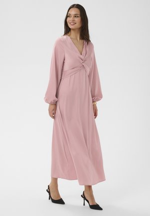 Femme portant une robe longue à manches longues de couleur rose poussiére avec un détail torsadé à l'avant, associée à des talons slingback noirs, debout et souriante.