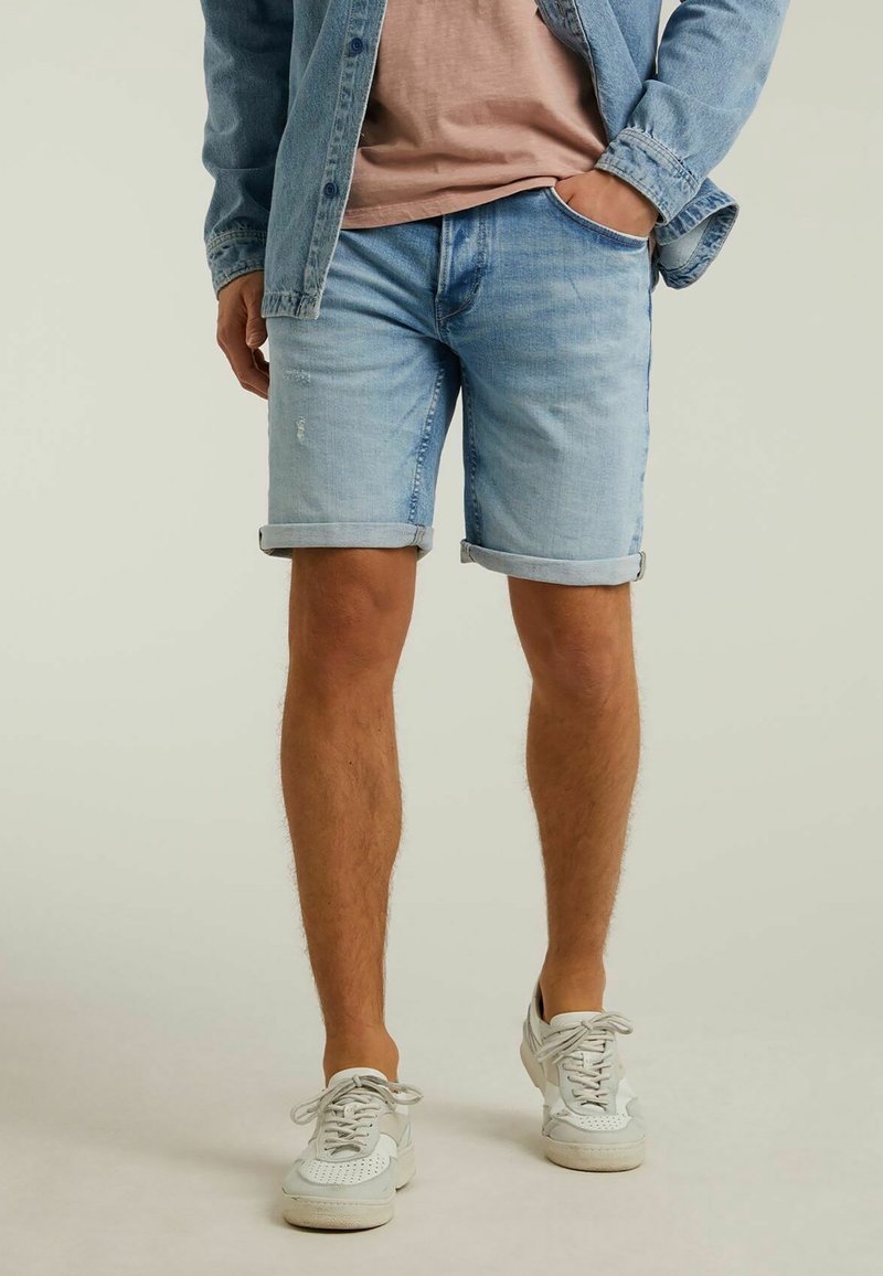 Homme portant un short en jean bleu clair retroussé, des baskets blanches, un t-shirt rose et une veste en jean avec une main dans la poche.