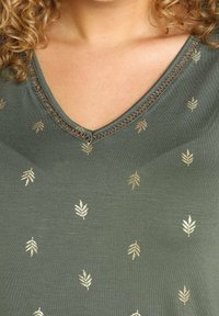 Grøn v-hals t-shirt lavet af blødt stof med guldfarvede bladmønstre og en dekorativt syet halsudskæring.