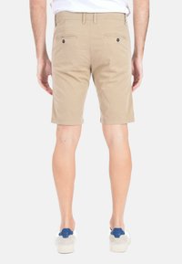 Pantaloni corti beige con una vestibilità classica, dotati di tasche posteriori con chiusura a bottone. Indossati con scarpe da ginnastica casual grigie e blu.