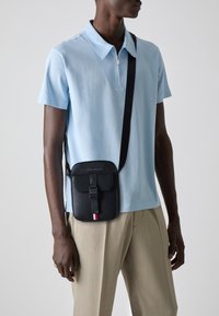Schwarze Umhängetasche mit strukturiertem Finish, ausgestattet mit einem Schnallenverschluss und einem gestreiften Akzent. Getragen über einem hellblauen Poloshirt.