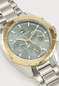 Tommy Hilfiger óra aranyozott kerettel, zöld textúrált számlappal, ezüst színű karkötővel és három, jól látható jelölésekkel ellátott alszámlappal.