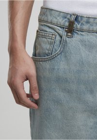 Close-up van de hand van een persoon die naast de voorkant van een lichtblauwe spijkerbroek en een wit geribd shirt rust.