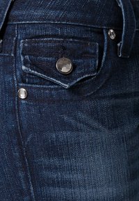 Mörkblå denimjeans med en framficka, silverfärgade metallknappar och synliga sömmar. Tyget har en lätt textur och en slät yta.