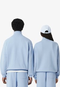 Sudaderas de color azul claro y de tallas grandes con cuellos altos, combinadas con pantalones a juego; la persona de la derecha lleva una gorra azul clara.