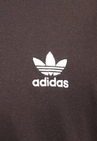 Ruda medvilninė marškinėliai su balta siūta Adidas logotipu ir trimis juostelėmis virš logotipo. Sklandžios tekstūros, paprasto dizaino.