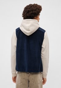 Gilet di velluto a coste blu navy con una superficie texture, abbinato a un hoodie beige chiaro, che mostra un design a strati e uno stile informale.
