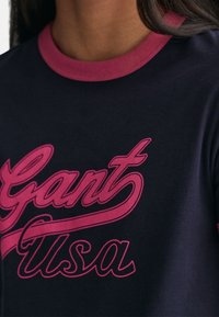 Camiseta de algodón azul marino oscura con un cuello circular rosa y gran texto rosa "Gant USA" en el pecho. Textura suave, diseño casual.