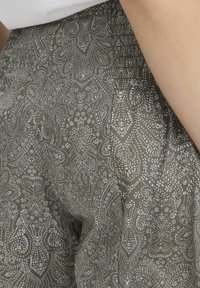 Gros plan sur un pantalon vert olive avec des motifs cachemire blancs complexes et des plis près de la taille, porté avec un haut blanc.