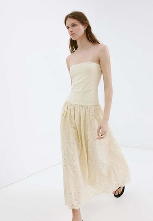Jeune femme portant une robe crème bustier avec jupe à œillets et sandales noires, marchant devant un fond blanc uni.