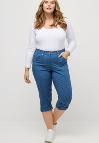Pantalons capri bleus en tissu lisse, avec des poches avant, des revers roulés et des coutures décoratives. Associés à un haut blanc à manches longues.