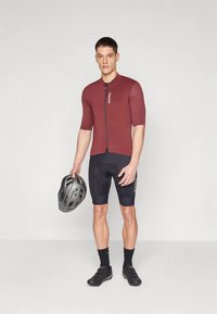 Maillot de cyclisme à manches courtes, couleur bordeaux, avec une fermeture éclair à l'avant, associé à un short rembourré noir. Le mannequin tient un casque de cyclisme noir.