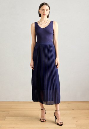 Robe plissée bleu marine avec un corsage côtelé, décolleté en V et jupe fluide. Assortie à des sandales à talons noires et un collier de perles.