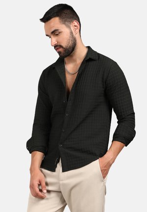 Camisa de botones de cuadros negros con una tela texturizada, mangas largas arremangadas hasta los codos, combinada con pantalones de color claro.
