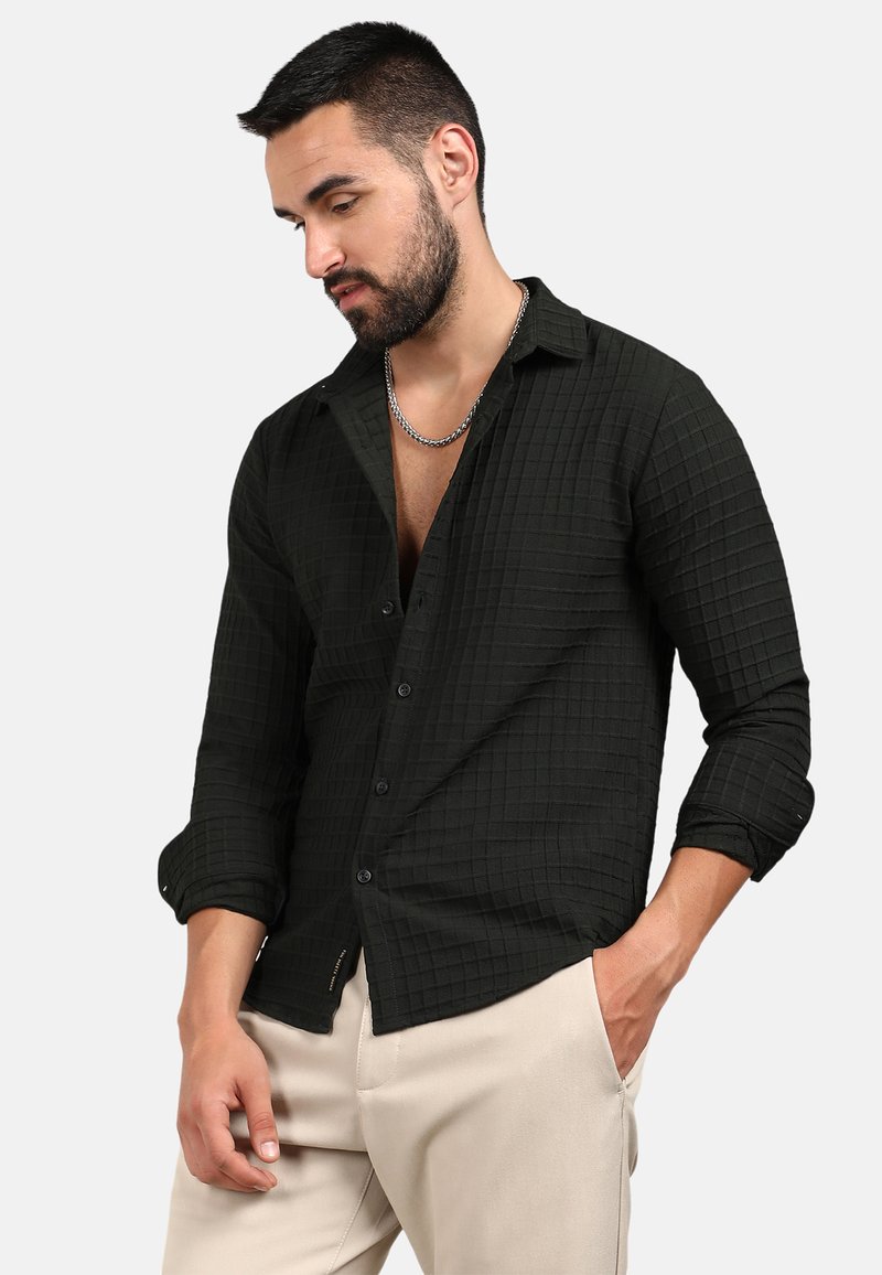 Camicia a quadri nera con tessuto testurizzato, maniche lunghe arrotolate fino ai gomiti, abbinata a pantaloni di colore chiaro.