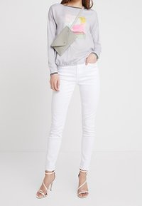 Graues Langarmoberteil mit pinkem und gelbem abstraktem Design, weiße enge Jeans und silberne hohe Sandalen mit Riemchen. Kleine graue Umhängetasche.