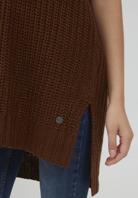 Pull en tricot marron présentant un motif texturé, des fentes sur les côtés et un petit logo métallique près de l'ourlet. Associé à un jean bleu.