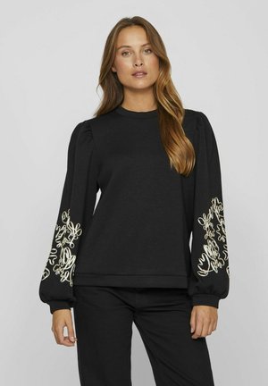 Sudadera negra con mangas abullonadas, que presenta un intrincado bordado en oro en los puños. Está hecha de una tela suave con un diseño de cuello redondo.
