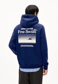 Persoon in een marineblauwe hoodie met de tekst "Open Water Free Swim" en een zwemmer graphic op de rug, gecombineerd met een gestreept shirt en donkere broek.
