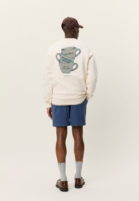 Ανοιχτό μπεζ φούτερ με γραφικό σχέδιο από στοιβαγμένες κούπες καφέ και κείμενο, σετ με μπλε σορτς και καφέ παπούτσια. Με υλικό από βαμβάκι και ριμπ μανσέτες.