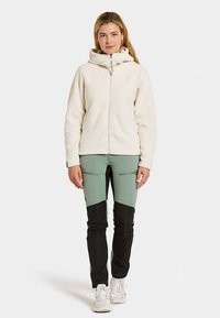 Kobieta stoi, ubrana w kremową polarową bluzę z zamkiem błyskawicznym oraz zielone i czarne spodnie w blokowane kolory, z białymi trampkami.