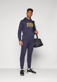 Marineblauwe sweatshirt en bijpassende broek met een gouden "PUMA sports club" logo. Model houdt een zwarte sporttas vast en draagt zwarte sneakers met groene accenten.