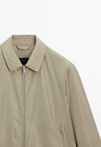 Béžová bunda na zip s špičatým límcem a kapsami na zip vpředu, zobrazena na bílém pozadí.