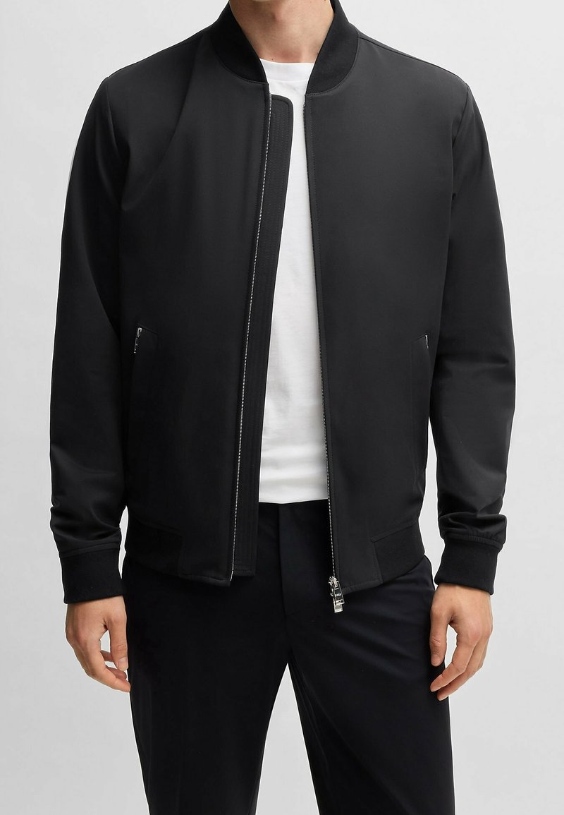 Homme portant une veste bomber noire zippée sur un t-shirt blanc avec un pantalon noir, debout devant un fond gris uni.