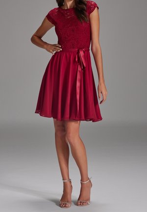 Frau trägt ein rotes Spitzenkleid mit einem Satinbandsgürtel und transparentem Rock, kombiniert mit silbernen High-Heel-Sandalen, steht auf grauem Hintergrund.
