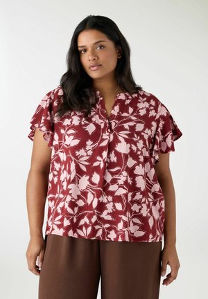 Donna che indossa una camicetta a maniche corte color marrone scuro con motivo floreale rosa e pantaloni larghi marroni, in piedi contro uno sfondo chiaro e semplice.