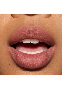 Gros plan sur des lèvres avec un rouge à lèvres mat nude. Les lèvres sont pleines, lisses et bien définies, avec une texture visible. Les dents sont partiellement exposées.