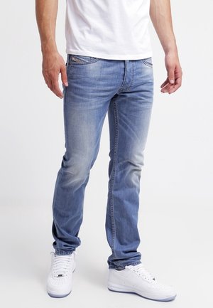 Mann trägt hellblaue, ausgewaschene Jeans, weiße Sneakers und ein weißes T-Shirt, steht vor einem einfarbigen weißen Hintergrund.