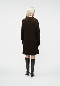 Frau mit langen blonden Haaren, die ein dunkelbraunes Strickkleid und schwarze, hochhackige Stiefel bis zum Knie trägt, von hinten auf einem weißen Hintergrund gesehen.