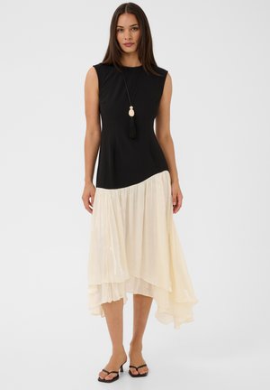 Femme portant un haut noir sans manches avec une jupe fluide asymétrique crème et des sandales noires à talons, debout devant un fond blanc uni.